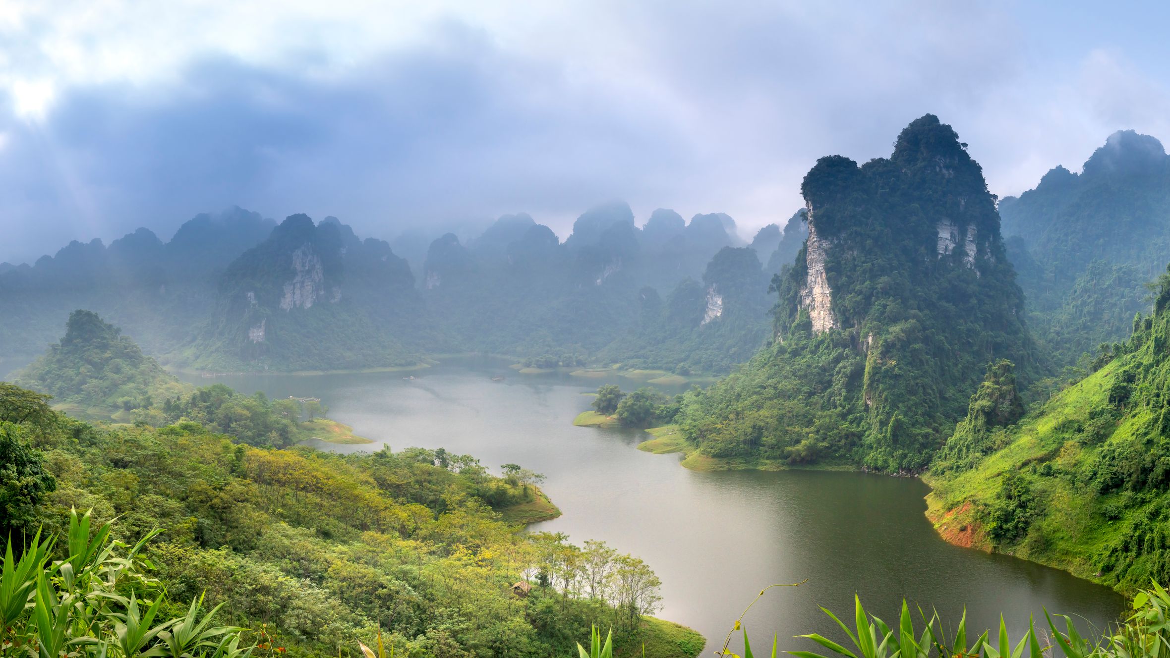 Wide angle of lake between high mounts with lush green trees and roadway under cloudy sky in misty weather