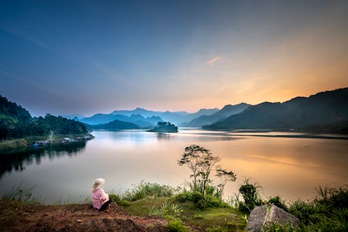 Back view of anonymous female tourist contemplating river and mount silhouettes in fog under cloudy sky at sundown