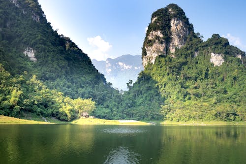 Scenic view of high mounts with lush green trees against rippled lake under cloudy blue sky in sunlight