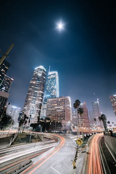 Long exposure of Los Angeles with skyscrapers and light trails under a full moon, capturing urban nightlife.