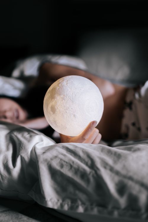 Free A woman lying in bed holding a glowing moon lamp in a cozy, dimly-lit setting. Stock Photo
