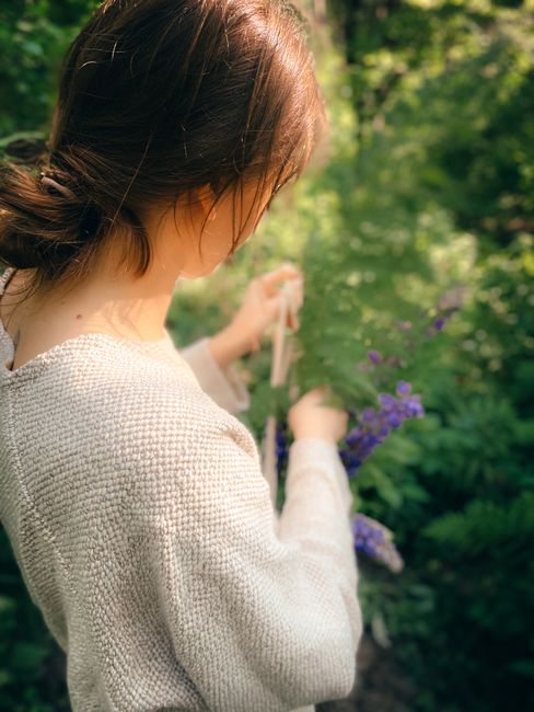 Side view of crop anonymous young woman with hair tied up collecting blooming violet flowers in summer garden on sunny day