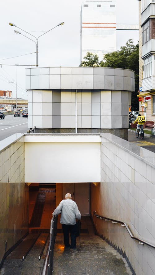 From above back view of crop anonymous aged man with cane walking down wet staircase of underground pedestrian passage in Russian city on cloudy day