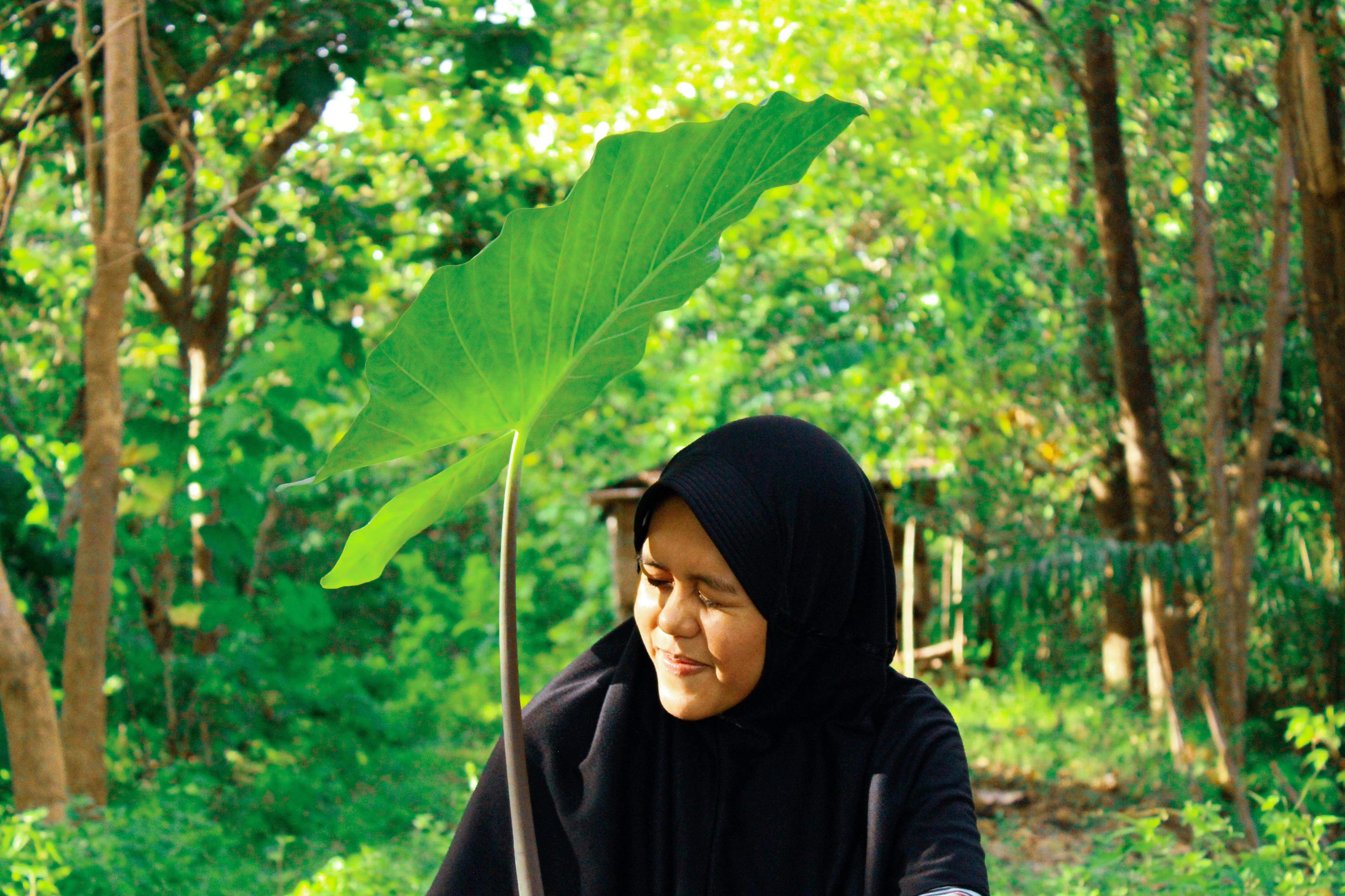 A woman wearing a hijab stands smiling among lush green trees, enjoying the outdoor scenery.
