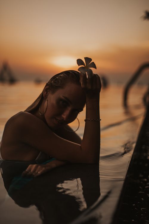 Free A serene portrait of a woman with a flower, enjoying a sunset in Bali's infinity pool. Stock Photo