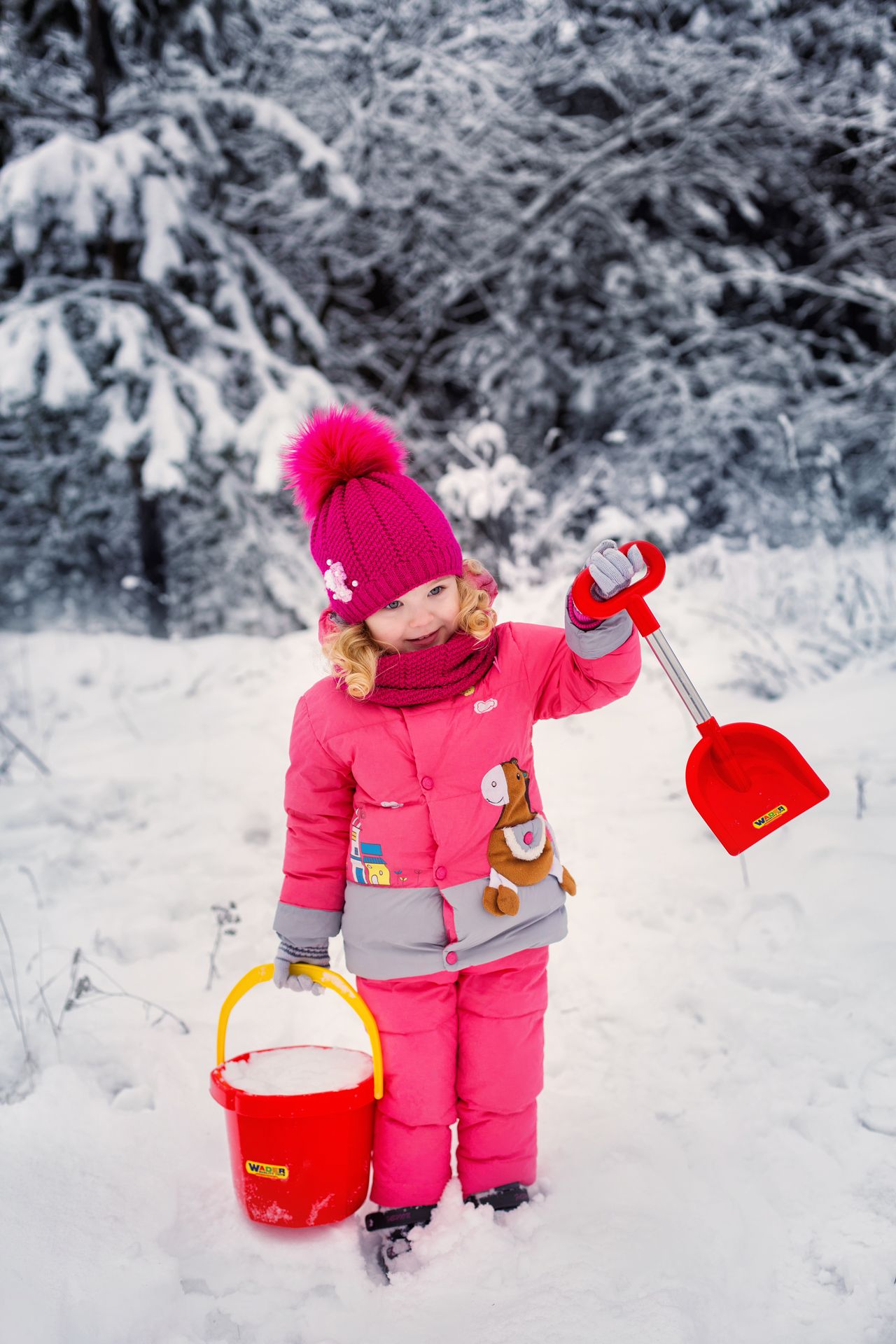 Little girl in a pink winter coat and snow pants holding a bucket and shovel in the snow