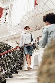 Full body smiling senior ethnic male in stylish summer clothes and hat leaning on stairway railing outside modern hotel building and looking at camera happily