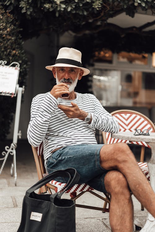Free Positive mature ethnic male in stylish summer wear and hat drinking aromatic coffee and looking at camera contentedly while sitting with legs crossed at table in outdoor cafeteria Stock Photo
