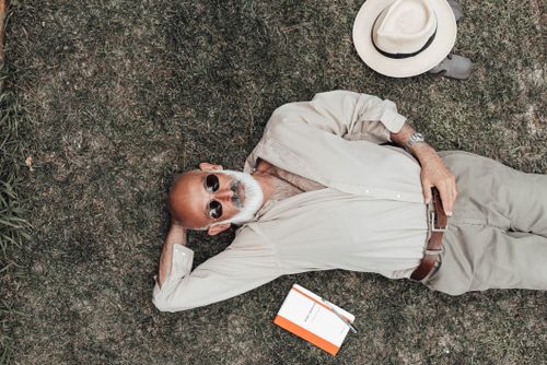 Free From above relaxed aged male in trendy wear and sunglasses lying on green grass near book and hat Stock Photo