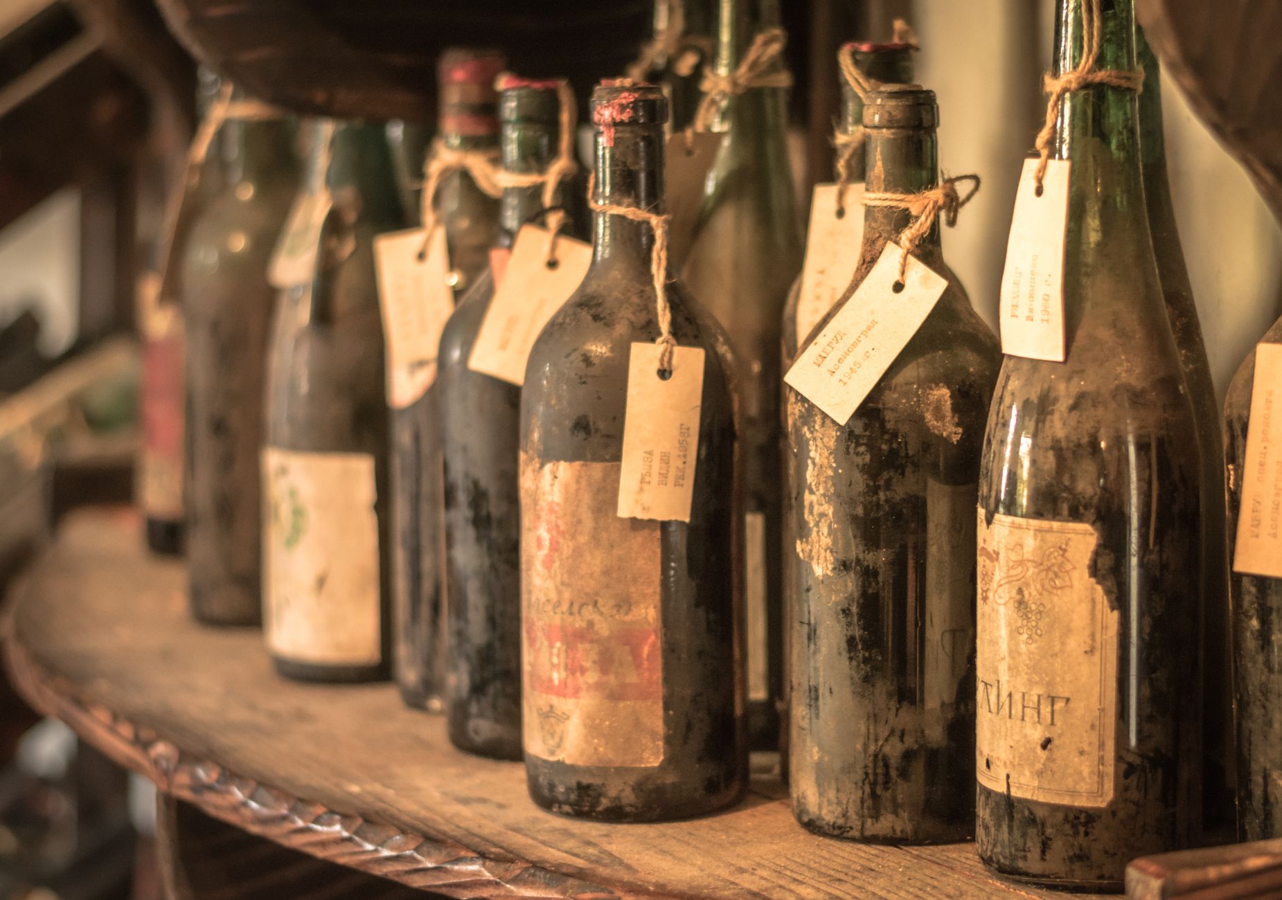 Close-up of old dusty wine bottles with tags on a wooden shelf in a vintage cellar setting.