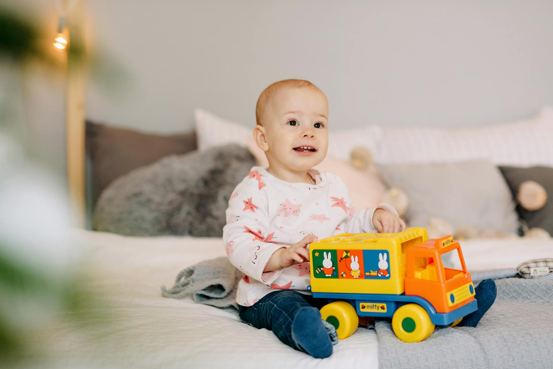 Happy baby boy sitting on bed playing with a colorful toy truck indoors.
