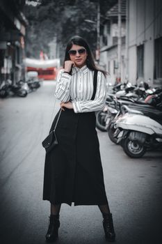Stylish young woman in black skirt and sunglasses standing confidently on a city street.