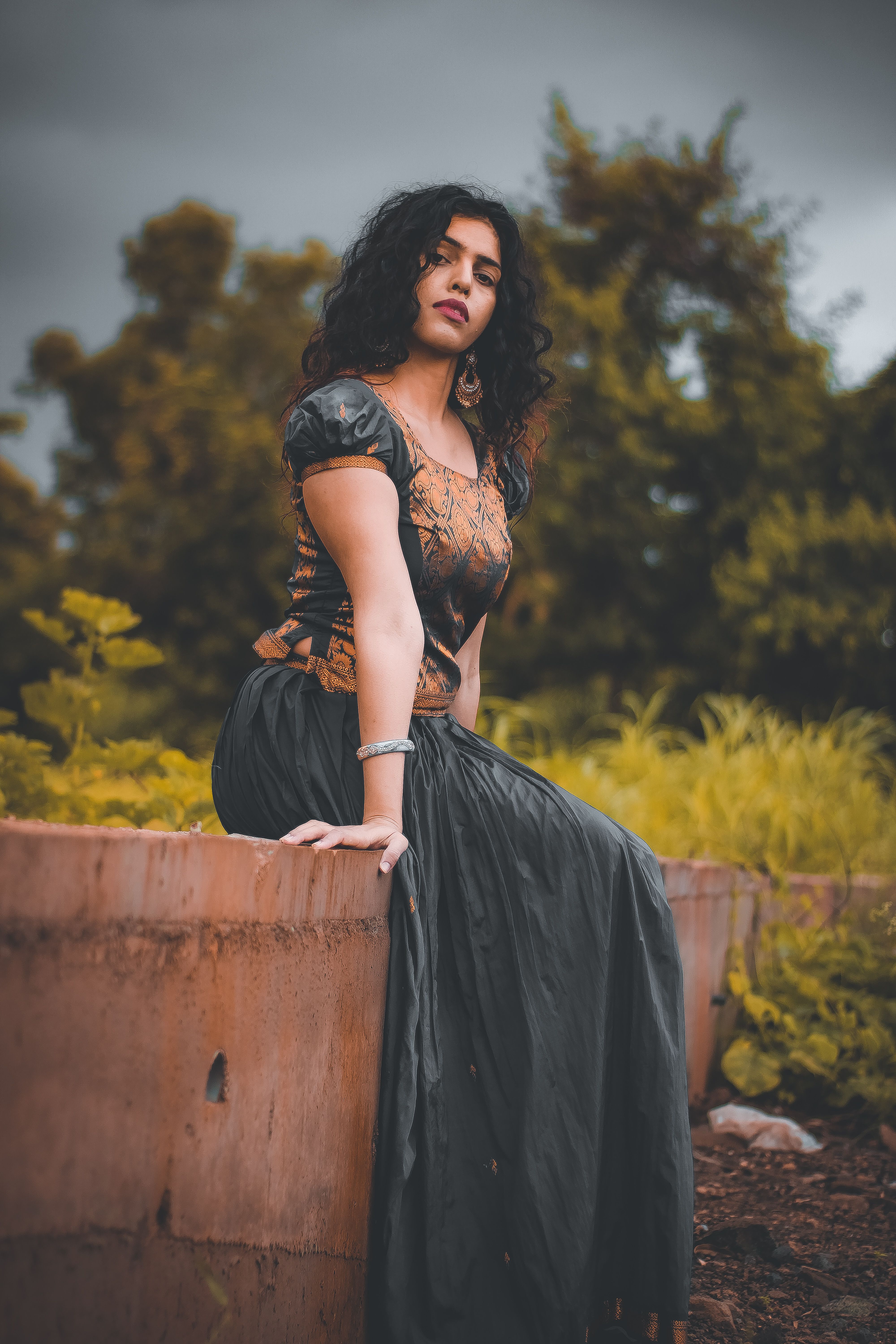 A woman with curly hair sitting gracefully near lush greenery outdoors.