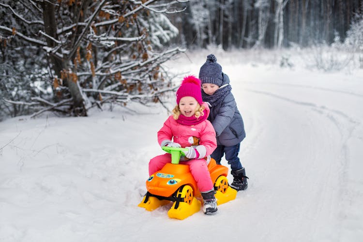 Boy In Blue Winter Clothes Pushing A Girl Riding On Toy Car On Snow Covered Ground