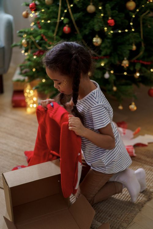 Free Young girl joyfully unboxes a Christmas sweater by the decorated tree, capturing holiday excitement. Stock Photo