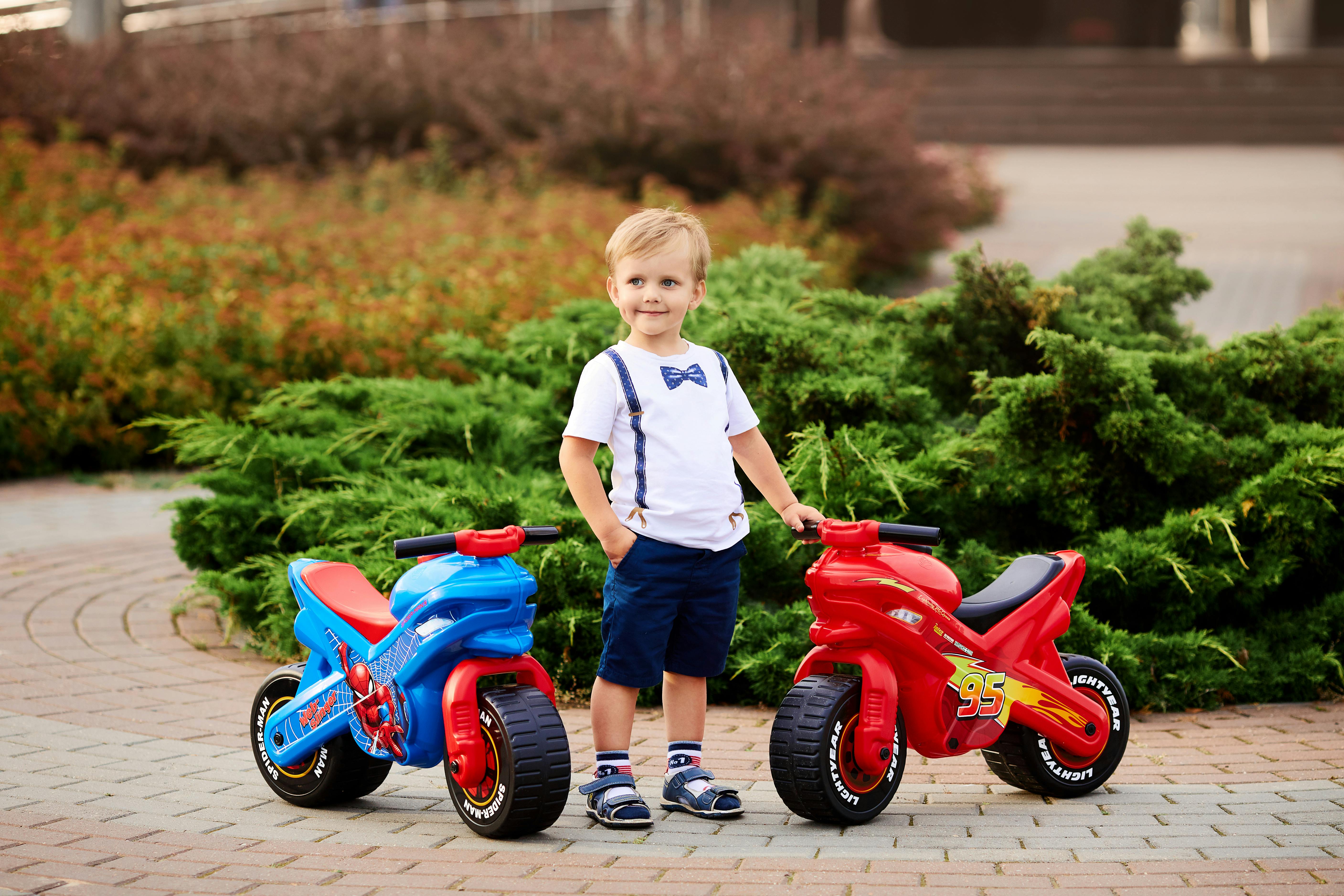 Smiling child standing between vibrant toy motorbikes outdoors in a park.