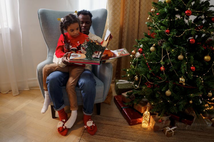 Dad And Daughter Reading A Fairy Tale Book While Sitting Beside A Christmas Tree