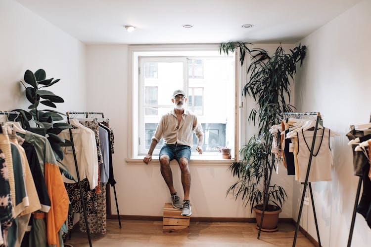 Senior Hipster Man On Windowsill In Clothes Shop