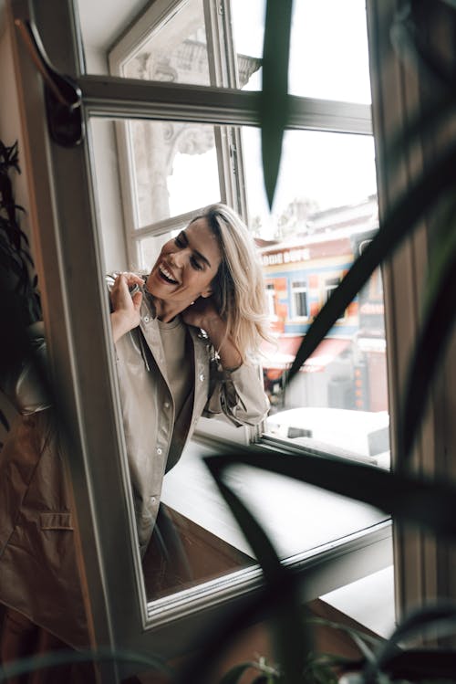 Adult happy female touching neck while looking away behind open window in building in daytime