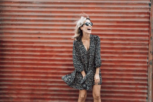 Cheerful female in stylish sunglasses and dress fluttering in wind laughing happily while standing against metal wall