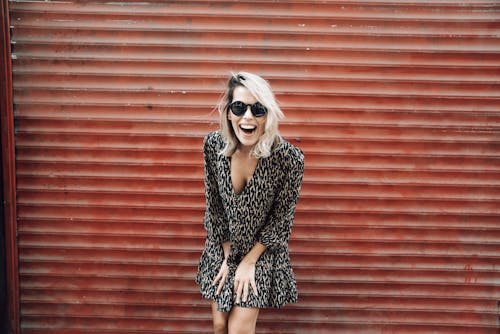 Happy female wearing stylish sunglasses and dress standing against red metal shutter and laughing widely
