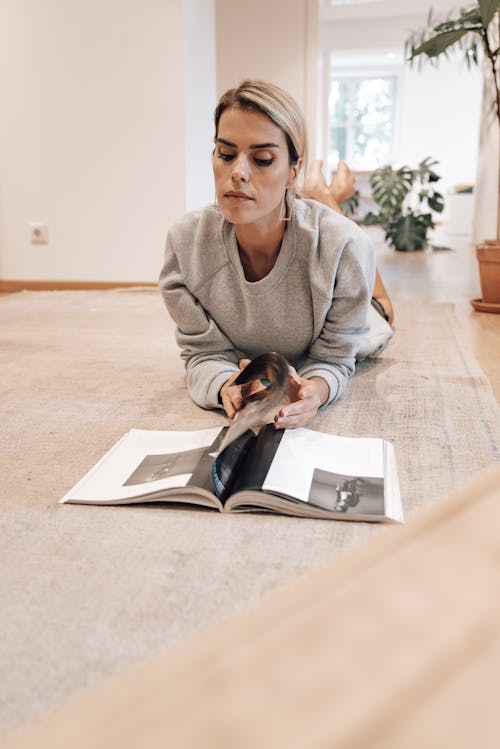 Serious female spending pastime at home and reading magazine while lying on floor in modern apartment