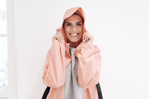 Free Happy young female wearing trendy rain coat with hood and smiling widely while standing against white wall Stock Photo