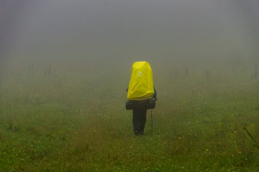 A lone hiker with a yellow backpack traverses a misty field, capturing a sense of adventure.