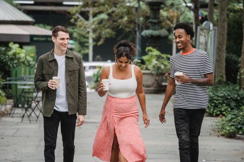 Free Cheerful young multiracial friends in stylish clothes smiling while walking together in city park with cups of takeaway coffee in hands Stock Photo