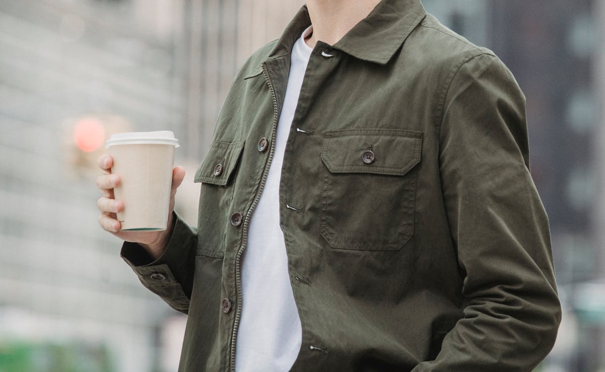 Free Side view of crop faceless young male in trendy outfit standing on modern city street with paper cup of coffee Stock Photo