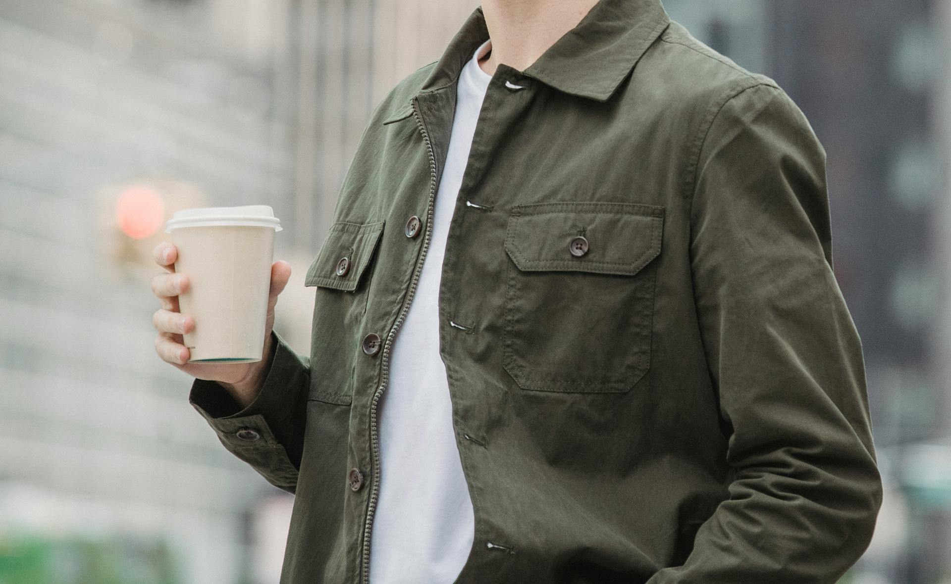 Side view of crop faceless young male in trendy outfit standing on modern city street with paper cup of coffee