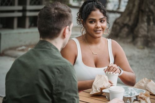Free Happy young Hispanic lady with dark curly hair in casual clothes drinking takeaway coffee and eating tasty dessert while spending time in park cafe with unrecognizable boyfriend Stock Photo