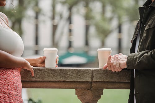 Free Side view of crop anonymous young couple in casual clothes standing on balcony with cups of coffee to go and chatting Stock Photo
