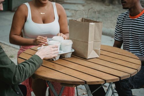 Free High angle of crop unrecognizable young diverse people sitting at wooden round table in outdoor cafe with takeaway drinks and paper bag Stock Photo