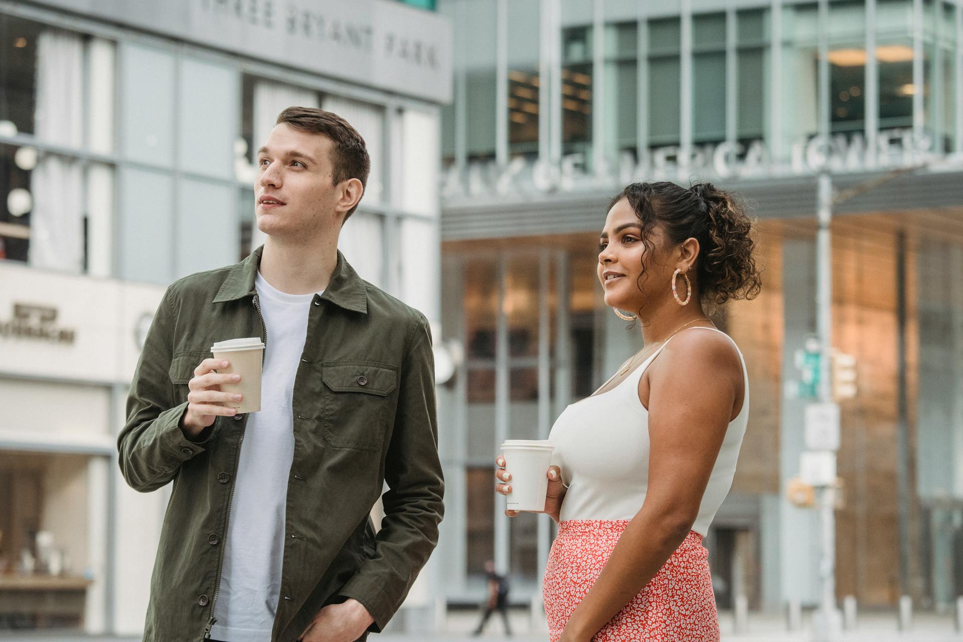 Positive young diverse coworkers in stylish clothes chatting on street near contemporary building and looking away while drinking coffee to go during break