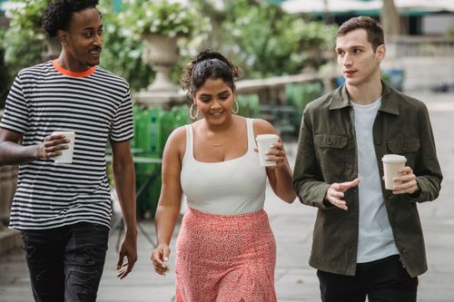Free Joyful young multiethnic students in casual clothes discussing lessons while walking together in park with takeaway cups of coffee Stock Photo