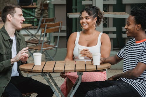 Free Cheerful young multiracial friends in casual clothes smiling while resting in outdoor cafe with cups of takeaway coffee and communicating Stock Photo