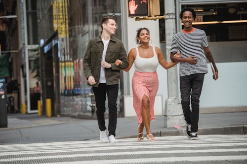 Full body of cheerful young Hispanic lady in stylish clothes holding hands of smiling diverse male friends while crossing road in modern city