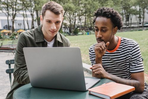 Free Concentrated young multiethnic male students sitting at table in campus park and browsing laptop while preparing for exams Stock Photo