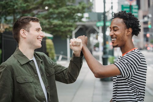 Side view of joyful young multiracial male friends in casual outfits smiling while bumping arms after meting on city street