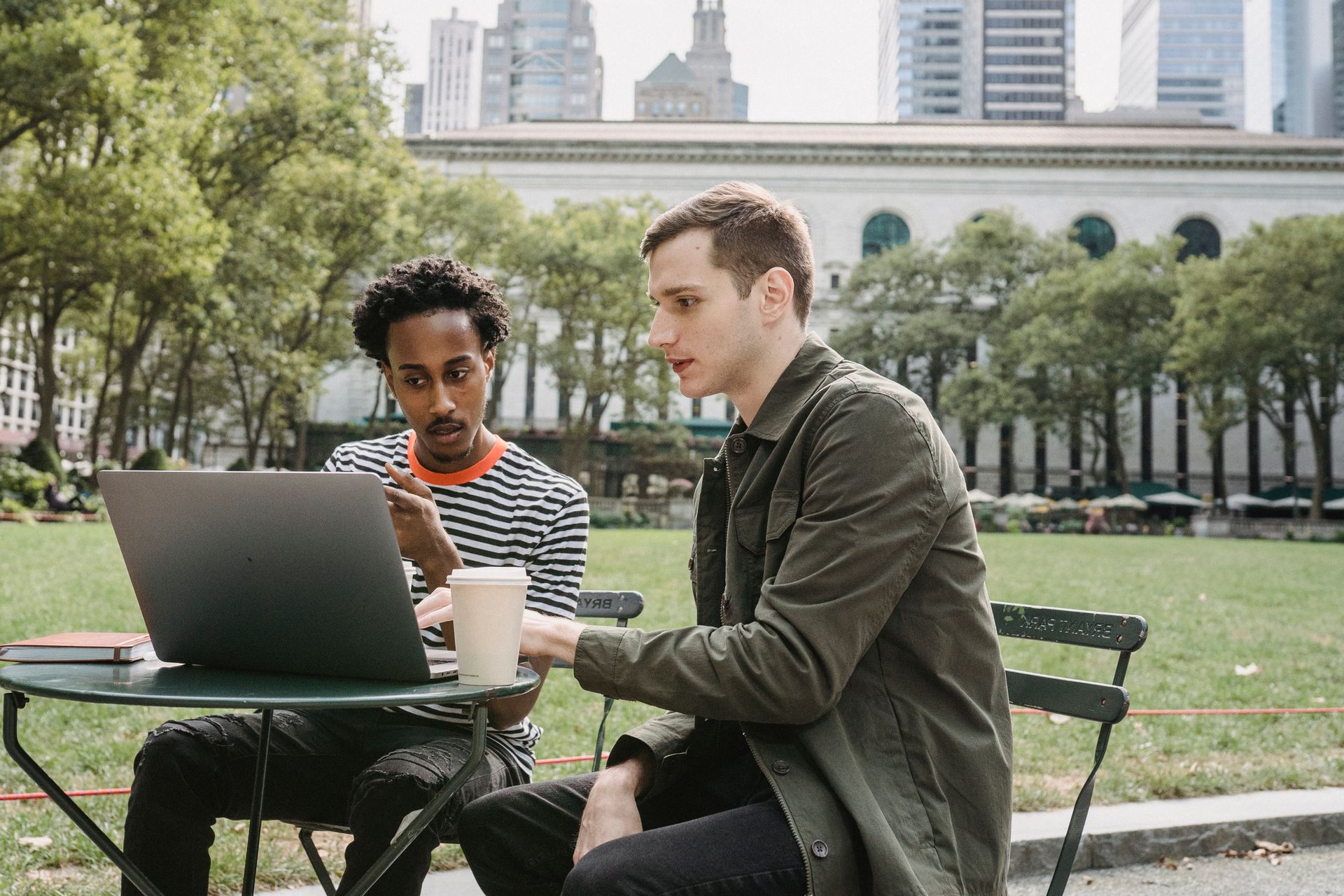 Focused young multiethnic male colleagues in casual clothes sitting at table in city park and working remotely on laptop