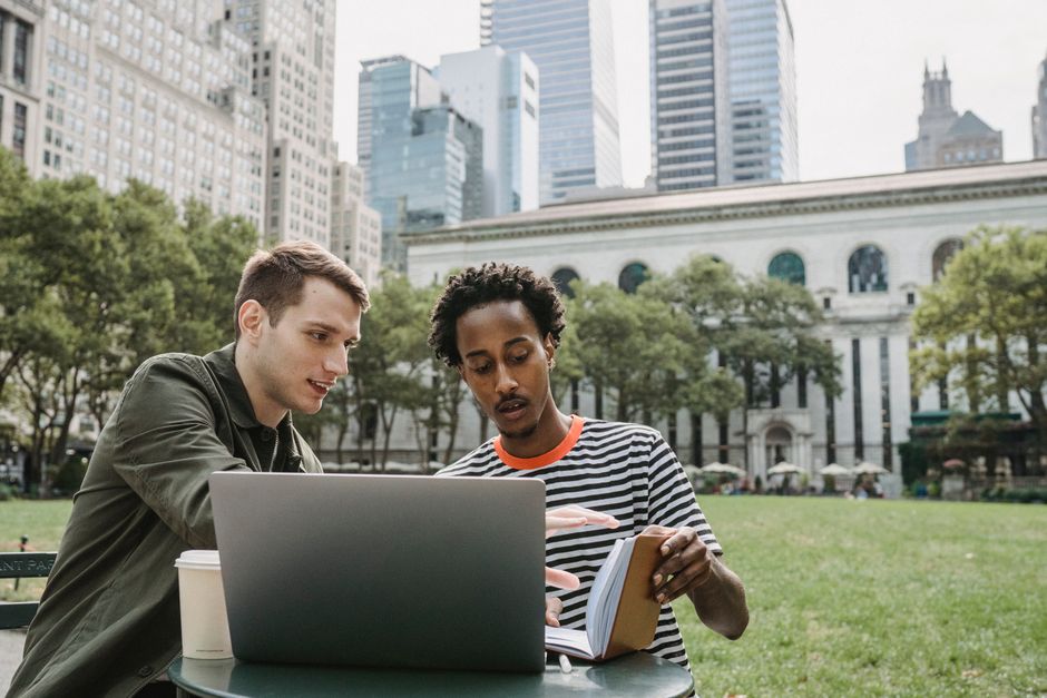 Serious young diverse male students reading notebook and using laptop during exam preparation together in campus park near modern skyscrapers Serious young diverse male students reading notebook and using laptop during exam preparation together in campus park near modern skyscrapers