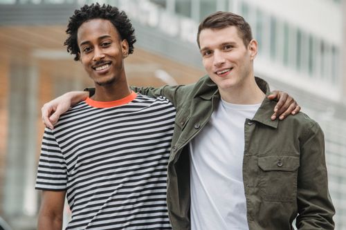 Free Content young multiethnic male friends in casual outfits smiling while standing on street with hands on shoulders and looking at camera Stock Photo