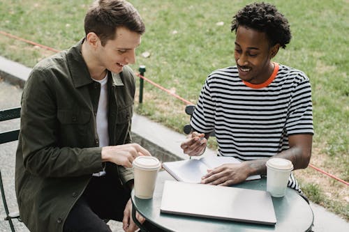 From above of cheerful multiracial male friends sitting at table with notebook and takeaway coffee while studying on university campus