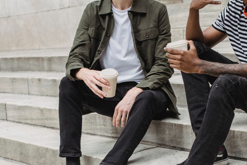 Unrecognizable multiracial male with paper cups of hot drinks sitting on concrete stairs of university building on street during coffee break Unrecognizable multiracial male with paper cups of hot drinks sitting on concrete stairs of university building on street during coffee break