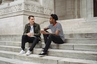 Full body of positive multiracial male students with takeaway coffee sitting on stone staircase of university building on street during break