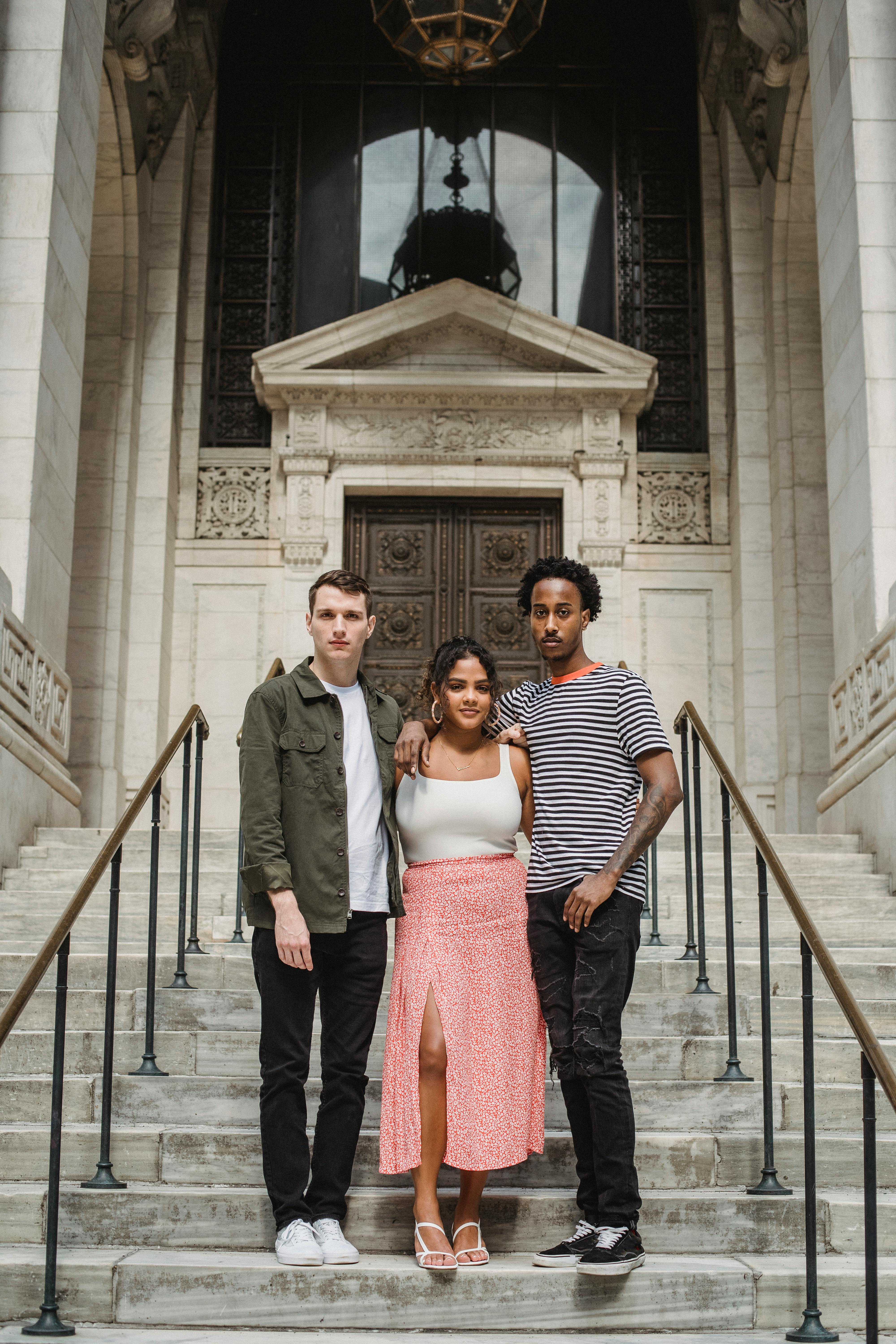 Full body of multiracial student looking at camera hugging while standing on stone staircase against aged building of university on street