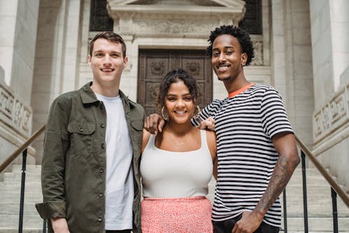 Cheerful young diverse best friends hugging and looking at camera while standing on aged building staircase