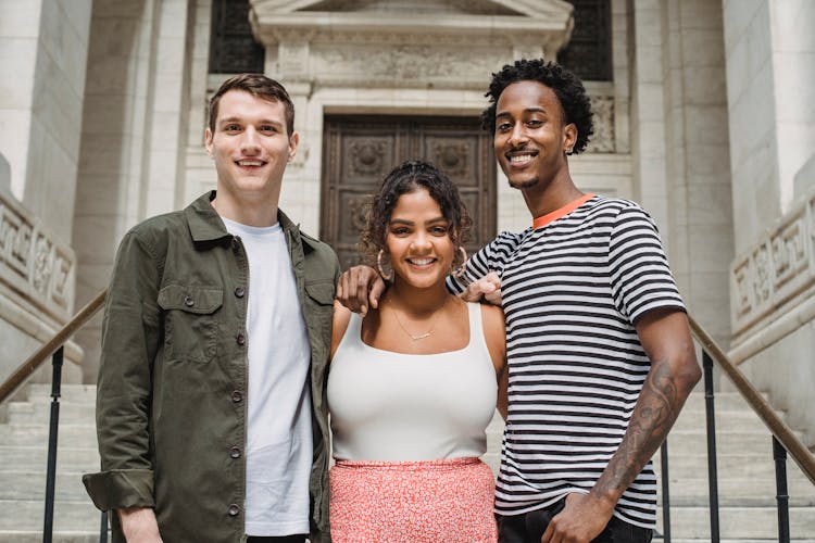 Positive Young Multiracial Students Hugging On Stairs Of Old University Building