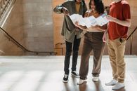 Concentrated young Hispanic lady reading map while standing near staircase of old building during vacation with crop multiethnic male friends